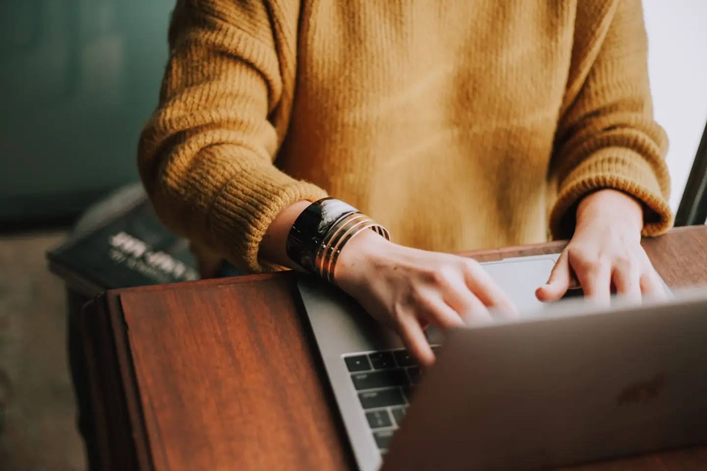 A person in a mustard sweater typing on a laptop at a wooden desk